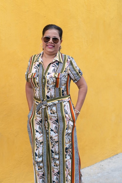 Mature woman in a dress and sunglasses stands against a yellow wall