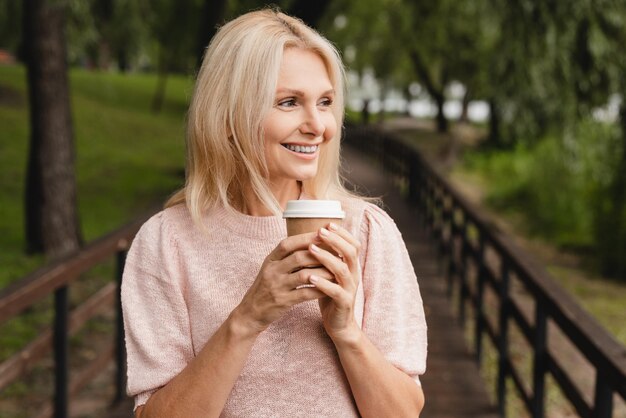 Mature woman drinking hot beverage coffee tea while walking in\
the forest park outdoors