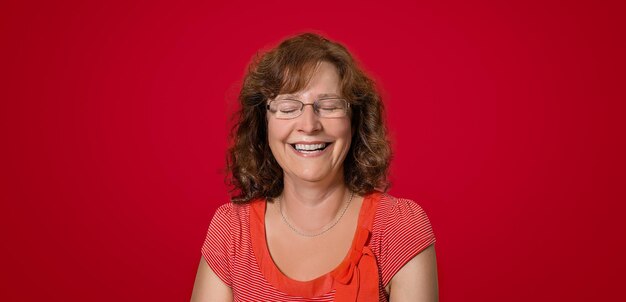 Mature woman in front of red background laugh and smile happy and joyful with closed eyes