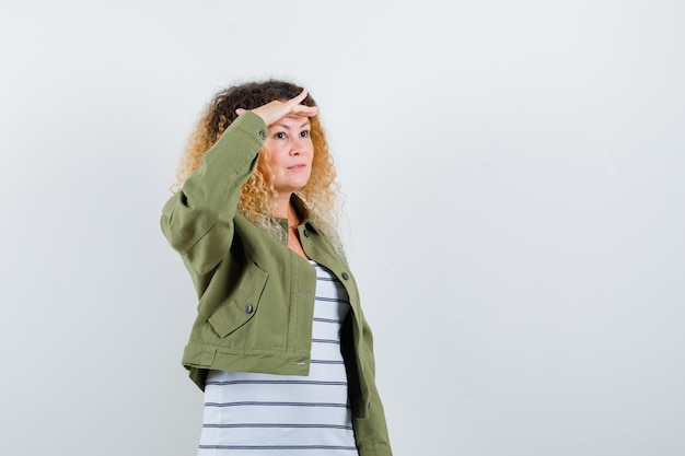 Mature woman in green jacket, t-shirt looking far away with hand over head and looking focused , front view.