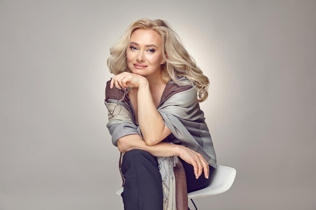 Mature woman in her fifties wearing in casual clothing and stylish plaid sitting over grey isolated