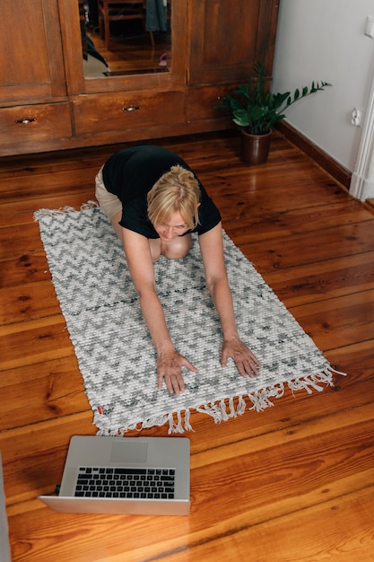 Зрелая женщина дома занимается спортивной йогой на полу с онлайн-занятиями на ноутбуке