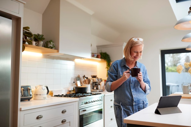 Зрелая женщина дома на кухне пьет кофе и смотрит на цифровой планшет