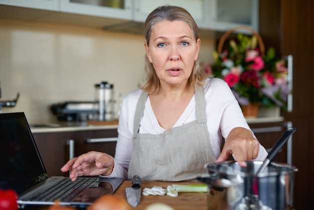 Mature woman in the kitchen prepares food and uses laptop