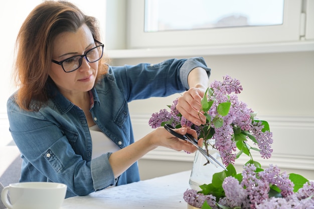 Mature woman making bouquet of lilac branches at home at table in living room