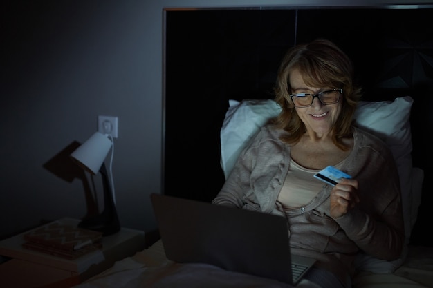 Mature woman paying with credit card online using laptop while she lying on her bed in the late evening