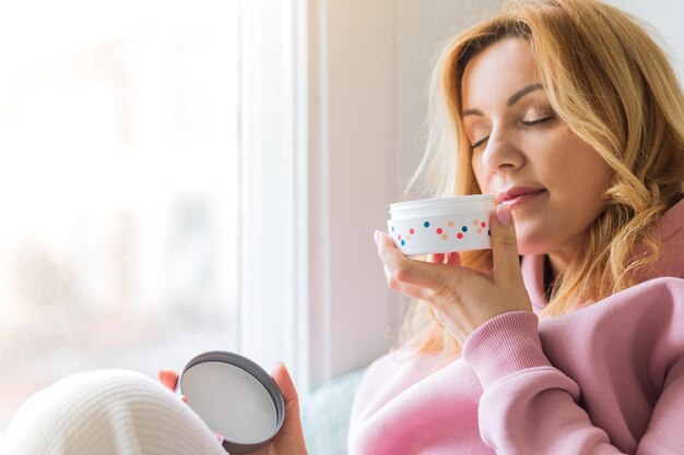 Photo a mature woman in a pink hoodie sits on the windowsill, holds a jar of cream in her hands and sniffs it.