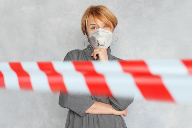 Mature woman in protective mask on gray background with warning tape