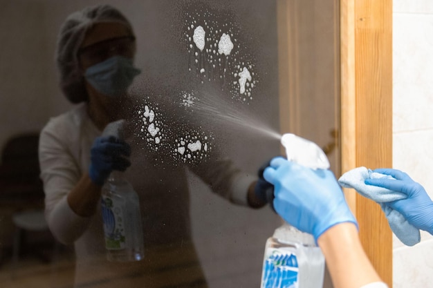 Mature woman in protective medical suit and respirator mask desinfecting furniture and cleaning door glass at hospital