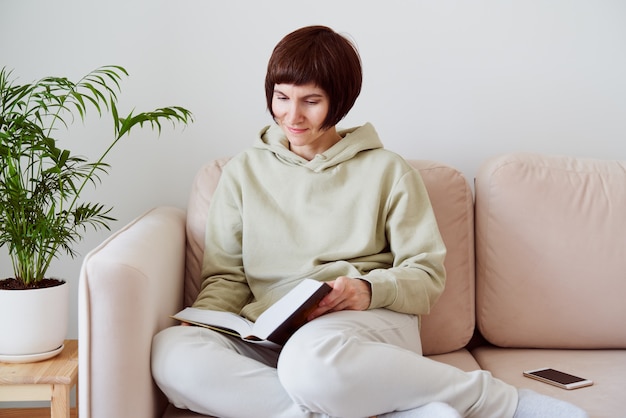 Mature woman reading book sitting on sofa thinking alone digital detox concept free time