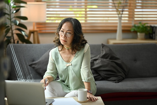 Mature woman reading news online checking social media on laptop computer while sitting on couch at home