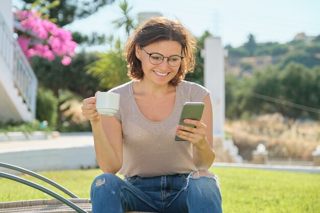 Mature woman resting outdoor, female sitting in rattan chair reading smartphone with cup of drink.