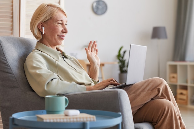Фото Зрелая женщина сидит в кресле с ноутбуком, глядя на монитор компьютера и машет подруге
