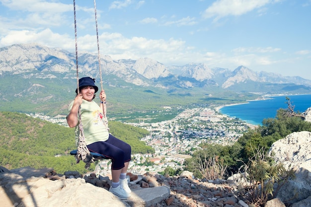 Mature woman in sportswear sitting on makeshift swing on a rope twine over an observation deck in the mountains