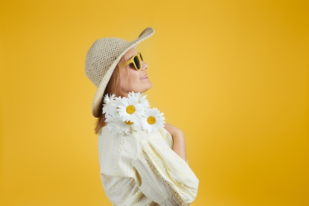 Mature woman in straw hat and sunglasses holds a bouquet of daisies isolate on bright yellow background