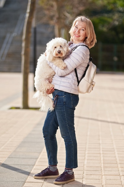 Mature woman walking with fluffy white dog in summer city.