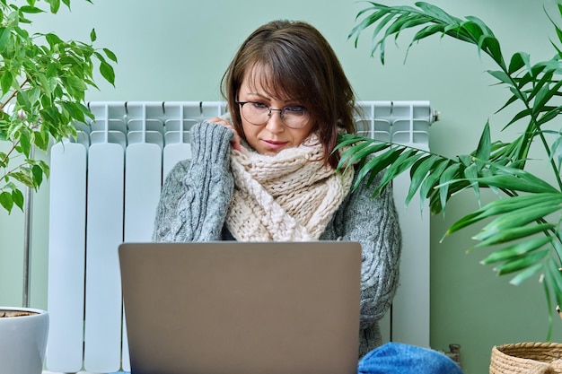 Mature woman in warm wool sweater scarf using laptop sitting near heating radiator at home Cold autumn winter season freelance work leisure lifestyle technology heat gas electric crisis energy saving