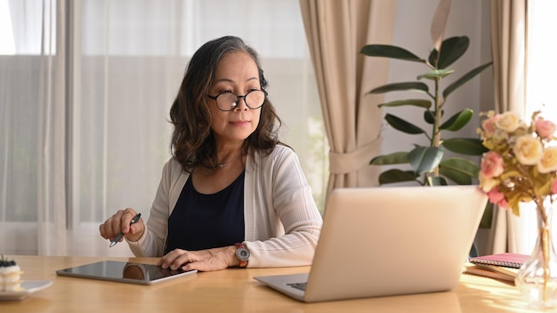 Donna matura che guarda il webinar online sul laptop e utilizza la tavoletta digitale durante il lavoro a distanza da casa