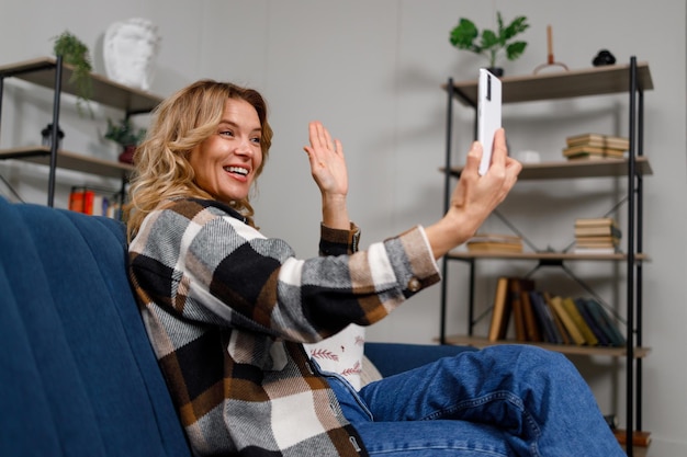 Mature woman waving hand during video call on smartphone while sitting on sofa