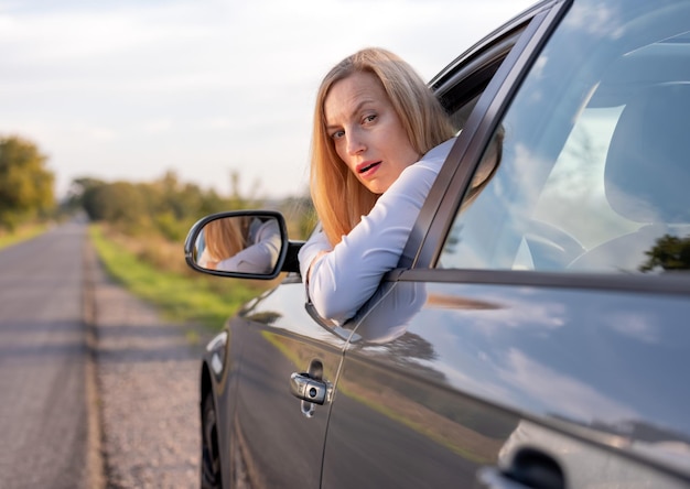 Фото Зрелая женщина со светлыми волосами смотрит из опущенного стекла современного автомобиля и чувствует себя потрясенной проблемы с транспортом на дороге проблемы во время вождения