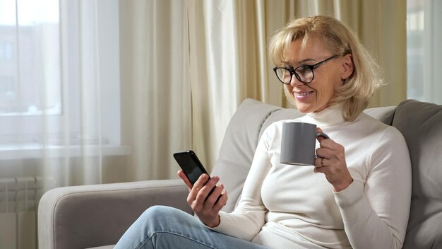 Mature woman with blonde hairstyle and in eyeglasses sips
coffee from mug and reads news online via phone sitting on sofa in
living room closeup