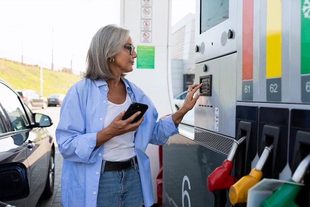 A mature woman with gray hair stands near a gas station and holds a smartphone