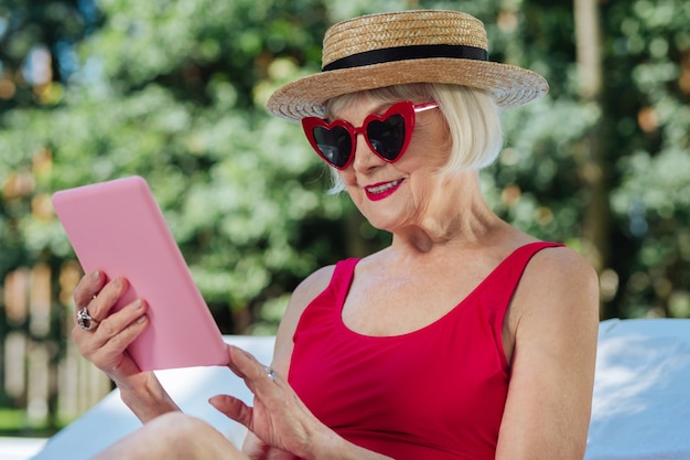 Зрелая женщина с красными губами чувствует себя отдохнувшей, читая электронную книгу на своем розовом ноутбуке