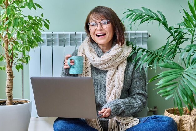 Mature woman in woolen clothes warming with cup of hot drink sitting near heating radiator smiling female looking at camera using laptop at home Heating season autumn winter lifestyle work freelance