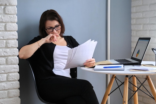 Donna matura che lavora in ufficio a casa, donna seduta alla scrivania con laptop leggendo documenti di finanze aziendali. lavoro a distanza, freelance, bollette domestiche, prestito bancario, informazioni sui pagamenti, spese domestiche