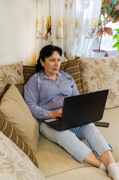 Mature woman works on laptop at home sitting on sofa in living room communicates shopping online
