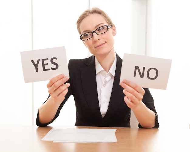 Photo maybe yes? thoughtful mature woman in formalwear sitting at the table and holding papers with yes and no on them