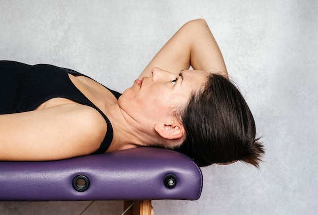 Фото Упражнение по методу маккензи для облегчения боли в шее, женщина лежит на массажном столе и опускает голову к полу, делая упражнения для снятия боли в шее.