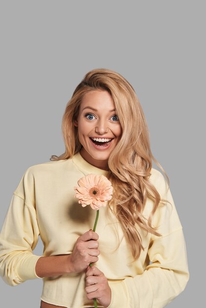 For me? surprised young smiling woman holding a flower and looking at camera while standing against grey background