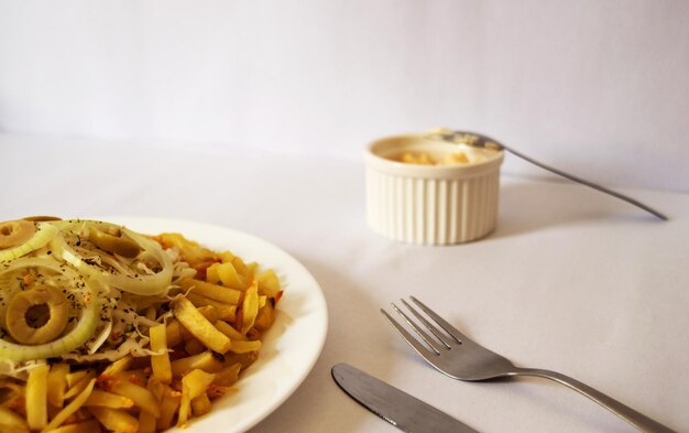 Meal on a white plate consisting of french fries and coleslaw\
with olives and onions with dehydrated seasoning placed on top of\
the potato and a container with mayonnaise next to it