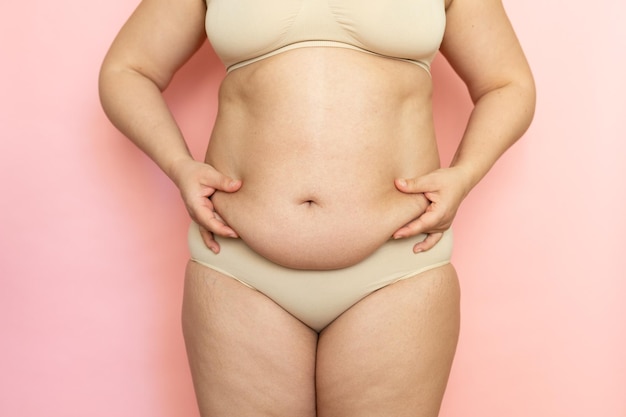 Measure woman sagging belly closeup folds on stomach loose skin\
and cellulite obesity naked overweight plus size girl on pink\
background in beige underwear concept of dieting and body\
control