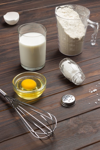 Мерный стакан с мукой, стакан молока, разбитое яйцо и соль, металлический венчик на столе. Темная деревянная поверхность. Вид сверху