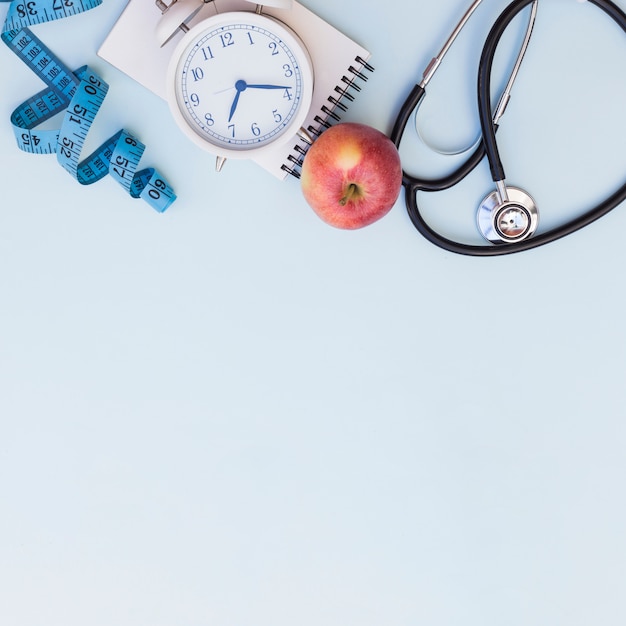 Measuring tape; alarm clock; spiral notepad; apple and stethoscope on blue background with copy space for writing the text