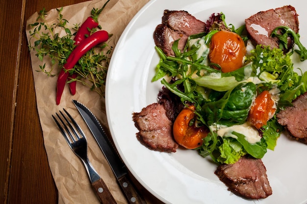 Meat beef steaks with greens, apricots and sauce on a large\
white plate on food mat - bamboo mat for food background. there are\
cutlery, a wooden pepper shaker and a salt shaker. barbeque,\
bbq