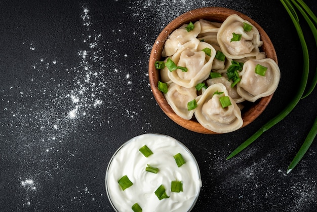 Meat dumplings in wooden bowl with sour cream and green onion on black background.