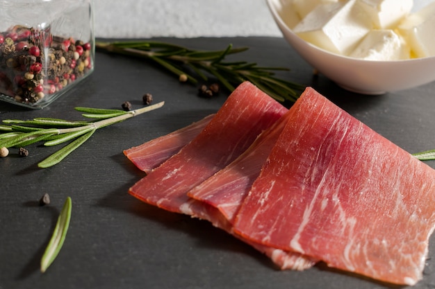 Photo meat plate, thin slices of prosciutto or spanish jamon with sprigs of rosemary, peppercorns and a plate with soft cheese on stone cutting board