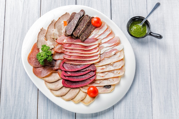 Фото Мясная тарелка с вкусными кусочками ломтики ветчины, помидорами черри, зеленью, мясом и соусом