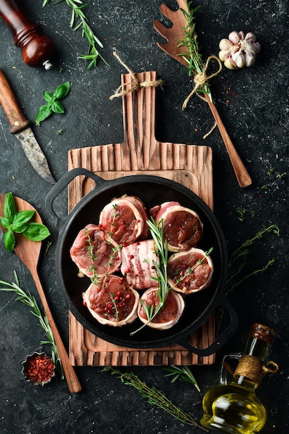 Foto carne carne cruda medaglioni di vitello avvolti nella pancetta disposti in una padella pronti per cucinare su uno sfondo di cemento nero vista dall'alto