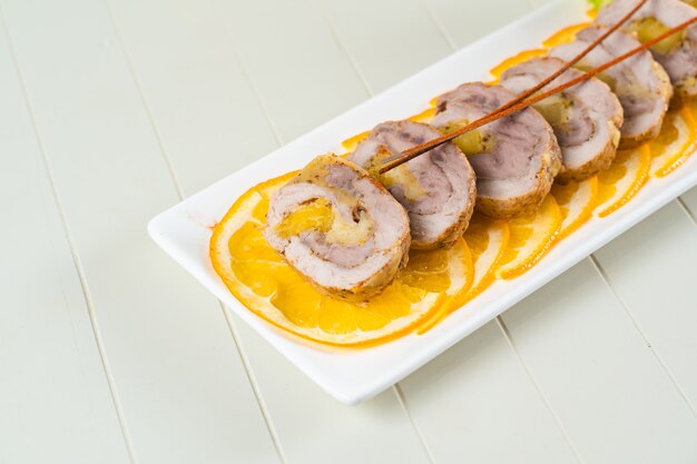 Meat rolls on orange slices on a rectangular white plate. Sliced pork appetizer.