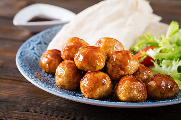 Meatballs in sweet and sour glaze on a plate with pita bread and vegetables in a Moroccan style on a wooden table.