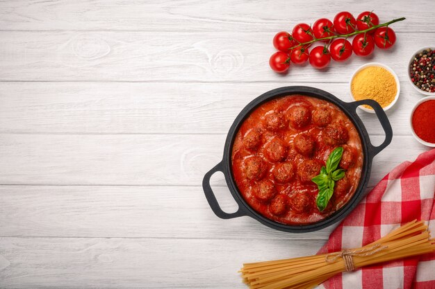 Meatballs in tomato sauce with spices and basil in a frying pan and cherry tomatoes on a white wooden board