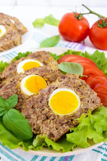 Meatloaf stuffed boiled egg with fresh tomato and lettuce leaves on a plate on a white wooden table. Close-up.