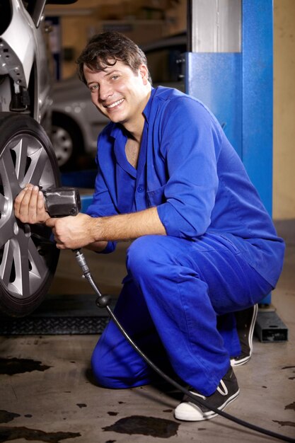 Foto meccanico e sorriso con la ruota in officina trapano elettrico e servizio auto o riparazioni per la manutenzione tecnico uomo e cambio pneumatici per cerchi automobilistici e ispezione del veicolo in ritratto