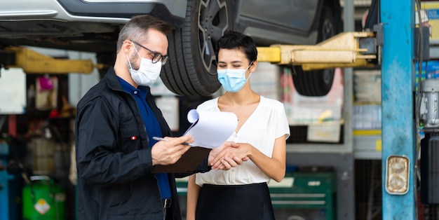 Механик мужчина и женщина-клиент носят медицинскую маску для защиты от коронавируса и проверяют состояние автомобиля перед доставкой.