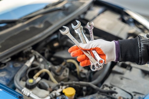 Mechanic\'s hand in a special glove holds a set of tools for the\
car near the car with an open hood. car repair concept