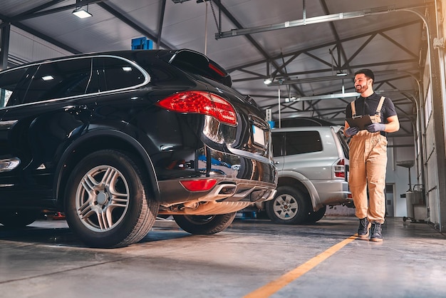 Mechanic writing on a clipboard at the repair garage while inspecting the car Full length